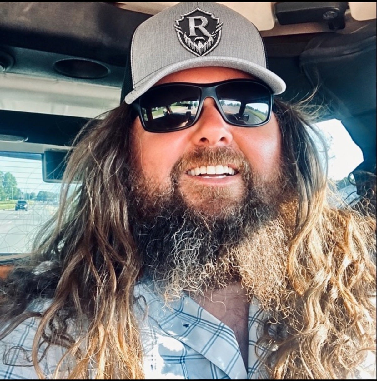 RuDaVoK off duty — long hair and beard, sunglasses, and his gray RuDaVoK shield hat, smiling in the truck